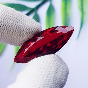 Può includere: Una pietra preziosa taglio marquise di colore rosso intenso e vibrante con una superficie sfaccettata che riflette la luce. La pietra è tenuta tra due dita coperte da guanti in tessuto bianco testurizzato.
