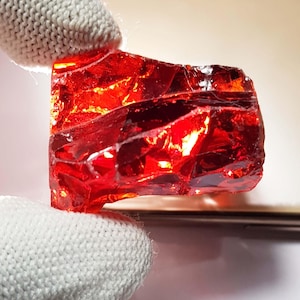 Könnte beinhalten: Ein leuchtender, durchscheinender, roter Edelstein mit einer rauen, unregelmäßigen Oberfläche. Der Stein wird von einer behandschuhten Hand gehalten, die seine feurige Farbe und kristalline Struktur hervorhebt. Der Stein scheint ein rohes, ungeschliffenes Mineral zu sein.