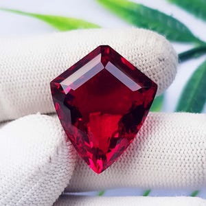 Puede incluir: Una piedra preciosa de color rojo intenso en forma de escudo con una superficie facetada. La piedra presenta una base puntiaguda y un borde superior plano, mostrando un color carmesí vibrante y translúcido con cortes geométricos nítidos.