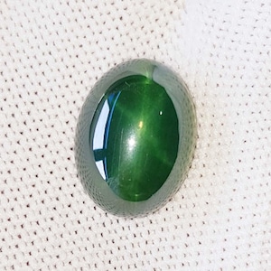 May include: A glossy, oval-cut gemstone in a rich green shade. The stone's surface is smooth and reflective, with a faint star-like pattern at its core. The gemstone is displayed on a white, textured fabric.
