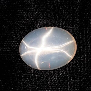 May include: Oval-shaped, translucent gemstone with a star-like pattern. The gem is a pale, milky white with a six-rayed star effect, set against a dark background. The star pattern is created by light reflecting within the stone.