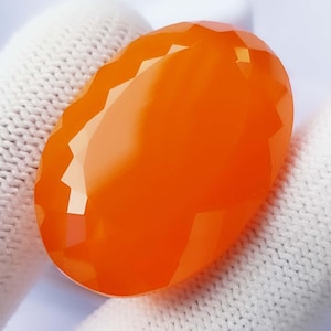 May include: A faceted, oval-shaped orange gemstone. The gem has a polished, glossy surface with multiple facets that reflect light. The color is a vibrant, saturated orange, and the overall shape is elongated. The gemstone is likely a cabochon or a similar cut.