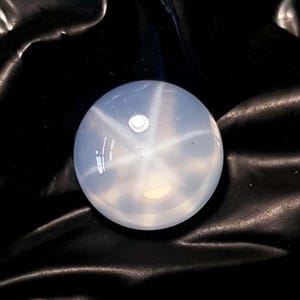 May include: A spherical, pale blue gemstone displaying a six-rayed star pattern. The gem is polished, reflecting light with a soft glow. The star effect is prominent, creating a captivating visual. The background is a dark, textured surface.