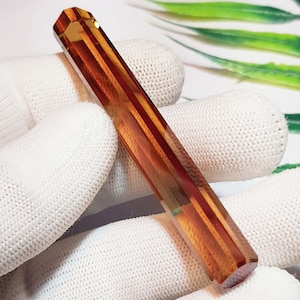 May include: A long, hexagonal-shaped rod made of translucent amber-colored material. The object features a smooth, polished surface with distinct linear facets that catch the light, creating a warm, glowing appearance throughout the length of the piece.