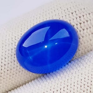 Puede incluir: Una vibrante gema azul de forma ovalada con un efecto de estrella. La superficie pulida refleja la luz, creando una exhibición cautivadora. La gema se encuentra sobre un fondo blanco texturizado, resaltando su color y brillo.