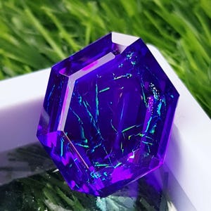 May include: A hexagonal gemstone with a deep purple colour and internal fractures. The fractures create iridescent streaks of blue and green. The gem is set against a white background, with green grass visible.