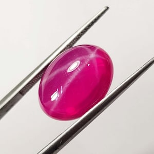 May include: A vibrant, oval-shaped ruby gemstone, held by silver tweezers. The polished stone displays a six-ray star effect, with a deep pink hue and a glossy surface. The gem is set against a plain white background.