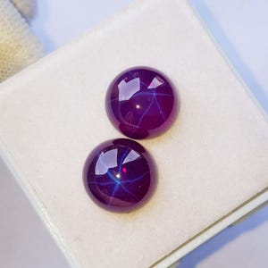 Könnte beinhalten: Zwei runde, im Cabochon-Schliff gearbeitete violette Edelsteine mit einem ausgeprägten sechsstrahligen Sterneffekt auf ihren polierten Oberflächen. Die Steine zeigen einen tiefen, gesättigten violetten Farbton mit Lichtreflexen auf den Kuppeln.