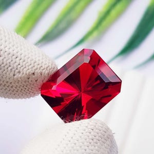 Può includere: Una gemma rossa dal taglio rettangolare con superfici sfaccettate che riflettono la luce. La pietra è tenuta tra due dita coperte da un tessuto bianco testurizzato, mettendo in risalto il suo colore cremisi intenso e la struttura geometrica.