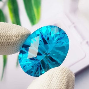 Op de afbeelding: Een levendige, ovaal geslepen edelsteen in een briljante cyaanblauwe kleur. De steen heeft ingewikkelde, geometrische facetten die een complex patroon van lichtreflectie en refractie creëren over het gepolijste, transparante oppervlak.