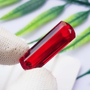 May include: A faceted, cylindrical gemstone with a deep, vibrant red color. The stone features a polished, translucent surface that catches the light, showcasing its elongated shape and precise geometric cuts along the edges and the rounded top end.