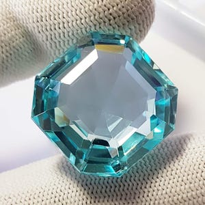 May include: A large, faceted gemstone with a light blue hue. The gem is cut into a unique octagonal shape with multiple facets that reflect light. The gemstone is held by a white glove.