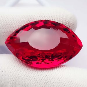 Könnte beinhalten: Ein Edelstein im Marquise-Schliff mit einer leuchtenden, tiefroten Farbe und einer facettierten Oberfläche, die das Licht reflektiert. Der Stein hat eine spitz zulaufende ovale Form mit symmetrischen Seiten, hoher Transparenz und Politur.