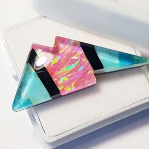 May include: A geometric resin pendant in a lightning bolt shape. The pendant features sections of pink, black, and turquoise, with iridescent accents. The pendant is displayed on a white surface.