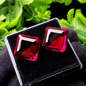 May include: Two square-cut, deep red gemstones with faceted surfaces are displayed side-by-side in a black velvet-lined display box. The stones exhibit a vibrant, translucent crimson hue with sharp, geometric edges and reflective facets.