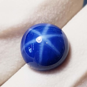 Puede incluir: Una gema pulida de color azul intenso con un prominente patrón de estrella de seis rayos. La piedra tallada en cabujón refleja la luz, creando un efecto de estrella en su superficie. La gema se encuentra sobre un fondo blanco roto.