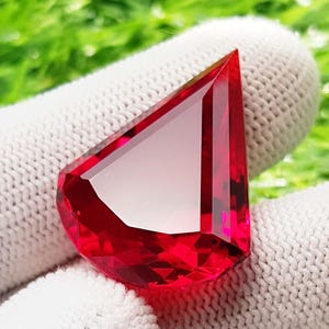 May include: A faceted, vibrant red gemstone with a unique geometric shape. The gem has a high degree of clarity, reflecting light and showcasing its deep color. The gemstone is set against a white background.