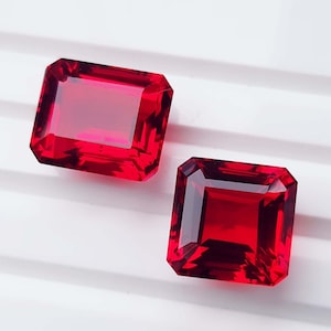 May include: Two faceted gemstones with a deep red color and a transparent finish. One stone features a rectangular cut, while the other stone is square-shaped. Both gems exhibit sharp edges and a polished surface that reflects light across their facets.