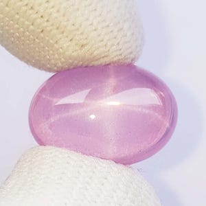 Puede incluir: Una gema redonda y pulida, de color púrpura claro, sostenida entre dos superficies de tela blanca. La gema tiene una superficie lisa y reflectante con reflejos internos. La apariencia general es un color púrpura suave y translúcido.