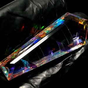 Puede incluir: Un gran cristal rectangular con iridiscencia de arcoíris. El cristal está facetado con múltiples planos, reflejando la luz en un espectro de colores. Los bordes son afilados y la forma general es geométrica.