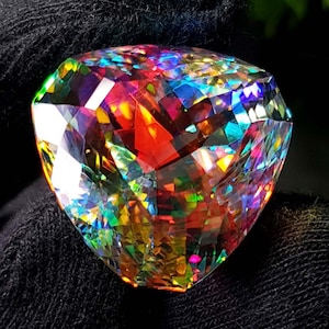 May include: A large, faceted gemstone with a complex, multi-colored internal structure. The gem displays a vibrant array of colors, including red, blue, green, and yellow, creating a dazzling display of light refraction. The gem is set against a dark background.