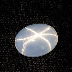 May include: An oval-shaped, pale blue gemstone with a six-rayed star pattern. The star effect is created by light reflecting off inclusions within the stone. The gem is set against a dark, textured background, highlighting its brilliance.