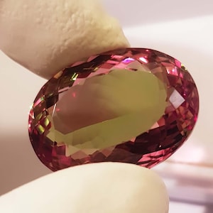 Könnte beinhalten: Ein oval geschliffener Edelstein mit einer Mischung aus rosa und gelben Farbtönen. Der Stein wird zwischen Fingern gehalten, wodurch seine facettierte Oberfläche und die Farbvariationen hervorgehoben werden. Die polierten Facetten reflektieren das Licht.