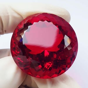 以下が含まれることがあります： 深紅色の大きな丸いファセット宝石。手袋をはめた手で持たれており、そのブリリアントカットと反射面が際立っています。宝石のファセットは、光と影の戯れを作り出しています。