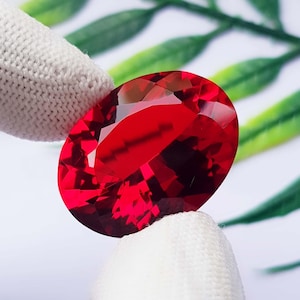 Può includere: Una gemma rossa dal taglio ovale con molteplici sfaccettature che riflettono la luce. La pietra è tenuta tra due superfici rivestite di tessuto bianco, mettendo in risalto il suo profondo colore cremisi, la chiarezza e la superficie lucida.