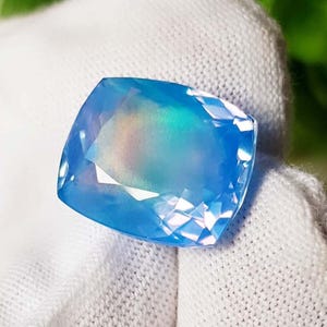 Può includere: Una gemma sfaccettata, taglio a cuscino, con una vibrante tonalità blu e lampi iridescenti di colore. La gemma è posta su uno sfondo bianco, mostrando la sua chiarezza e brillantezza. Le sfaccettature della gemma riflettono la luce.