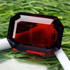 Può includere: Una gemma rettangolare sfaccettata di colore rosso intenso. La superficie lucida presenta bordi netti e definiti. La gemma è posta su uno sfondo di erba verde, evidenziando il suo colore e la sua chiarezza. Il taglio della pietra è a taglio smeraldo.