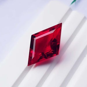 Puede incluir: Una piedra preciosa de color rojo intenso con facetas, cortada en una forma afilada y alargada de diamante o cometa. La piedra presenta múltiples superficies pulidas que reflejan la luz, creando profundidad y claridad interna en el color.