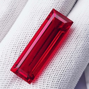 May include: A vibrant, rectangular-cut, red gemstone. The gem has a polished surface with sharp, angled facets, reflecting light. The gemstone is displayed on a white, textured fabric background.