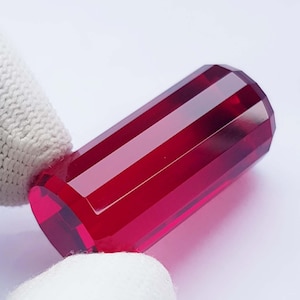 May include: A faceted, elongated ruby gemstone. The gem is a deep, rich red color with a polished surface. The shape is rectangular with angled sides and ends. The gemstone is held by a white glove.