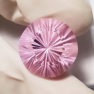 Può includere: Una gemma rosa rotonda e sfaccettata. La gemma presenta un motivo radiale di tagli, creando un effetto a stella. Il colore è un rosa tenue e chiaro. La gemma sembra essere tenuta da una mano guantata. Per gioielli.