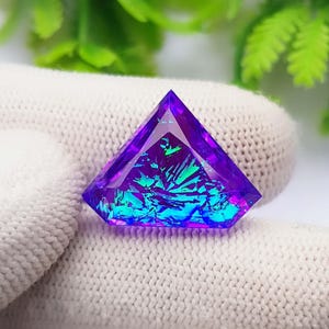 May include: A triangular-cut gemstone featuring a deep purple outer border and a vibrant, iridescent center with shifting blue and green light reflections. The stone has a faceted surface that creates a sharp, geometric shape.