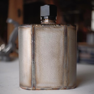 Puede incluir: Una petaca metálica rectangular hecha a mano con marcas de soldadura visibles. La petaca tiene un acabado plateado mate y una tapa negra. La petaca está diseñada con bordes redondeados y un cuerpo cilíndrico.
