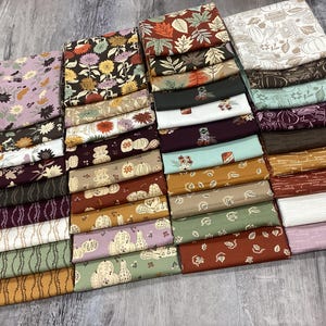 May include: A collection of folded fabric squares featuring various autumn-themed patterns, including pumpkins, fall leaves, sunflowers, and floral motifs in a palette of orange, brown, green, cream, purple, and white, arranged in a grid-like display.