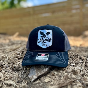 May include: A black and gray trucker hat with a white patch featuring a stylized eagle and the word "Merica" above "1776". The hat has a curved brim and a mesh back.
