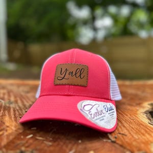 May include: A bright pink trucker hat with a white mesh back. The hat features a brown leather patch with the word "Y'all" stitched on it. The brim is curved, and the hat has a tag that says "Evolve Elude".