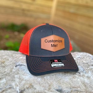 May include: A gray and orange Richardson 112 trucker hat with a leather patch that says "Customize Me!". The hat has a black brim and a mesh back. The hat is sitting on a rock.