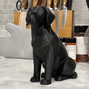 May include: A black geometric dog sculpture in a seated position. The dog has a faceted design and is placed on a white countertop. Kitchen knives in a wooden block are in the background.