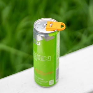 May include: A green and silver aluminum can with the word "GREEN" printed on it. An orange can opener is attached to the top of the can. The background is a blurred green.