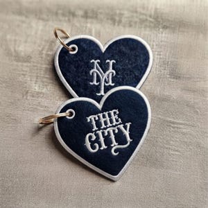 May include: Two heart-shaped dark navy blue felt keychains with white embroidered edges. The top heart features the white embroidered letters NY, while the bottom heart displays the white embroidered text THE CITY. Each has a gold metal ring.