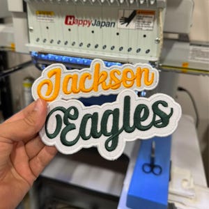 May include: Two embroidered patches with white borders and a scalloped edge. The top patch features the word Jackson in yellow cursive script. The bottom patch features the word eagles in dark green cursive script on a white background.