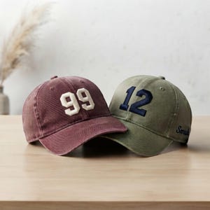 May include: A maroon baseball cap with the number 99 in white felt-style lettering and an olive green baseball cap with the number 12 in navy blue felt-style lettering, featuring the name Sarah embroidered in navy blue on the side panel.
