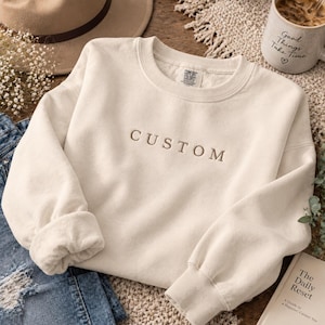 May include: A cream-colored crewneck sweatshirt featuring the word CUSTOM embroidered in a dark brown serif font across the chest. The garment has a ribbed collar, cuffs, and hem, with a Comfort Colors brand label visible inside the neckline.