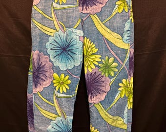 May include: Blue floral print pants with a vibrant pattern of large flowers in shades of purple, yellow, and turquoise. The pants are hanging on a metal hanger against a dark background. The floral design is repeated throughout the fabric.