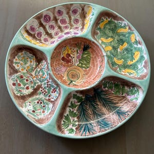 May include: A round, divided serving platter with a light green rim and six sections. Each section features a different hand-painted garden scene with flowers, vegetables, and foliage in shades of brown, green, yellow, and pink. The center has a woven basket design.