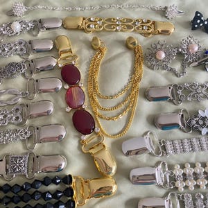 May include: A collection of vintage belt clips and accessories in various designs. The clips feature silver and gold tones, with embellishments like rhinestones, pearls, and decorative patterns. Some clips have chain accents and oval-shaped red stones.